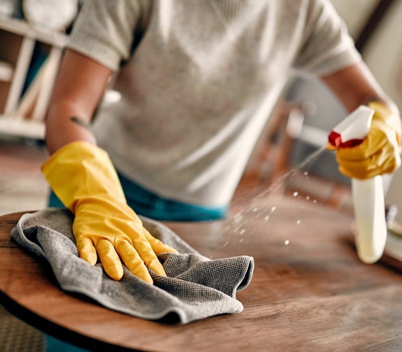 Person wearing yellow gloves cleaning a wooden table with a cloth and spray bottle.