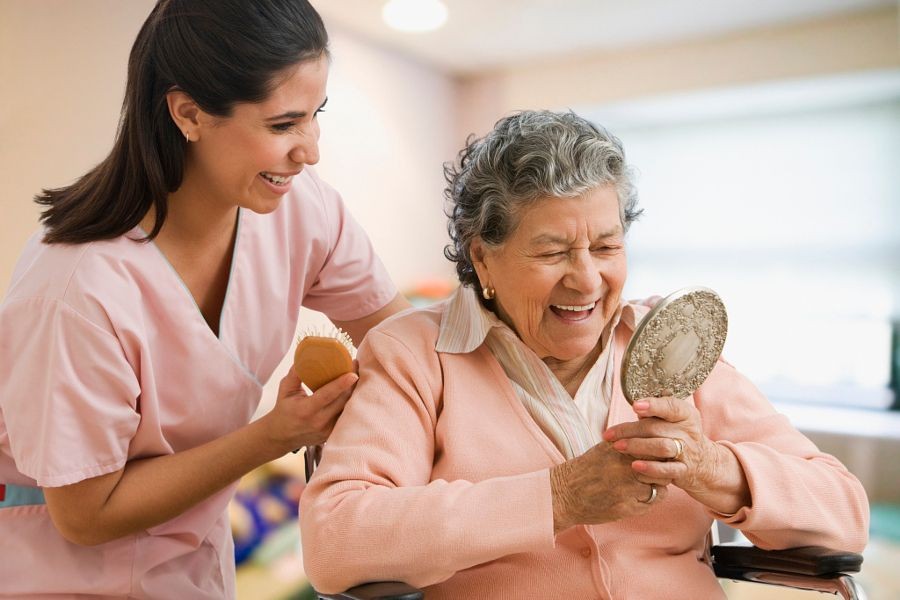 Caregiver assisting an elderly person holding a mirror and a brush in a well-lit room.
